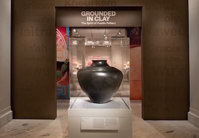 Large, beautifully proportioned clay pot on a pedestal in a museum gallery under a sign that reads "Grounded in Clay: The Spirit of Pueblo Pottery"