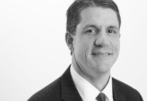 Professional head-and-shoulders portrait of a smiling man in a dark suit and tie in a studio setting.